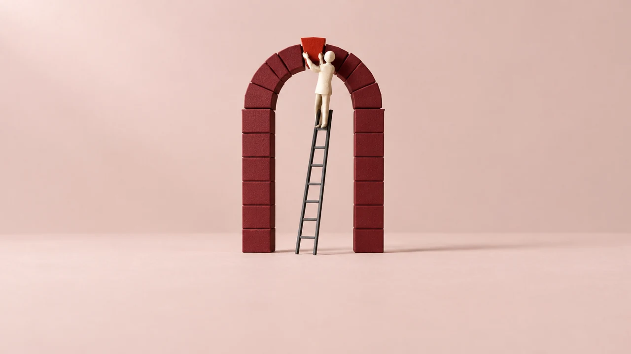 A small faceless figure on a ladder sets a red keystone into the crown of a tall paper arch