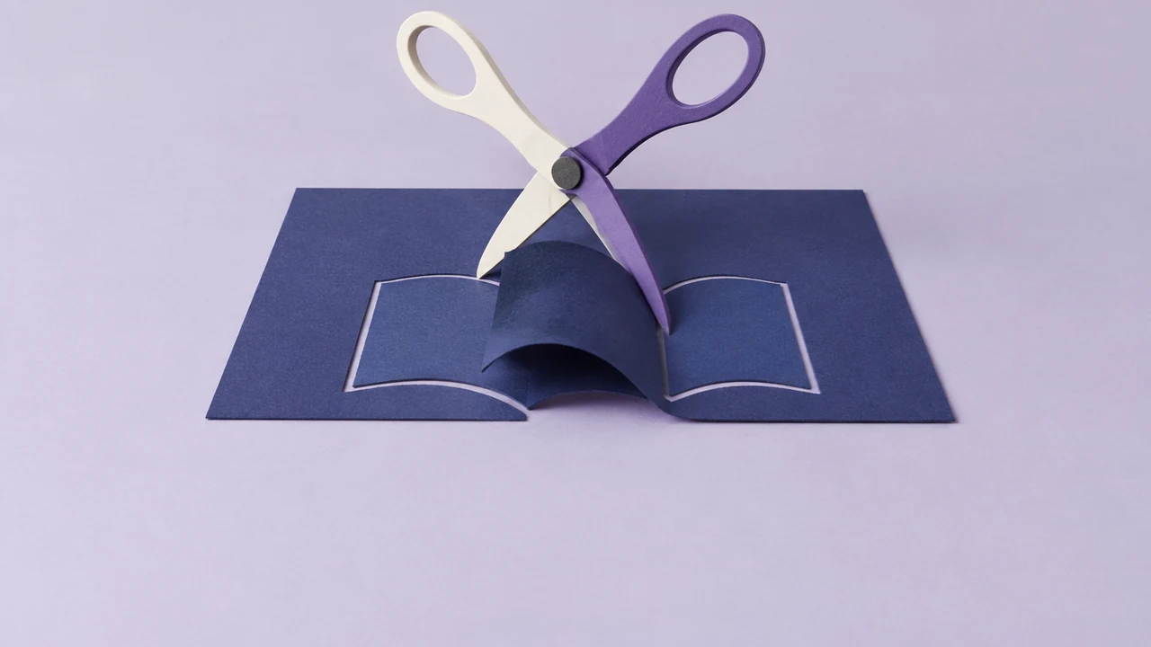 A large pair of scissors, one blade white and one violet, halfway through cutting an open book shape from a sheet of card